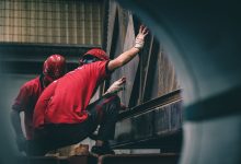 Dois trabalhadores usando capacetes vermelhos montam componentes metálicos dentro de uma fábrica.