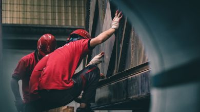 Dois trabalhadores usando capacetes vermelhos montam componentes metálicos dentro de uma fábrica.