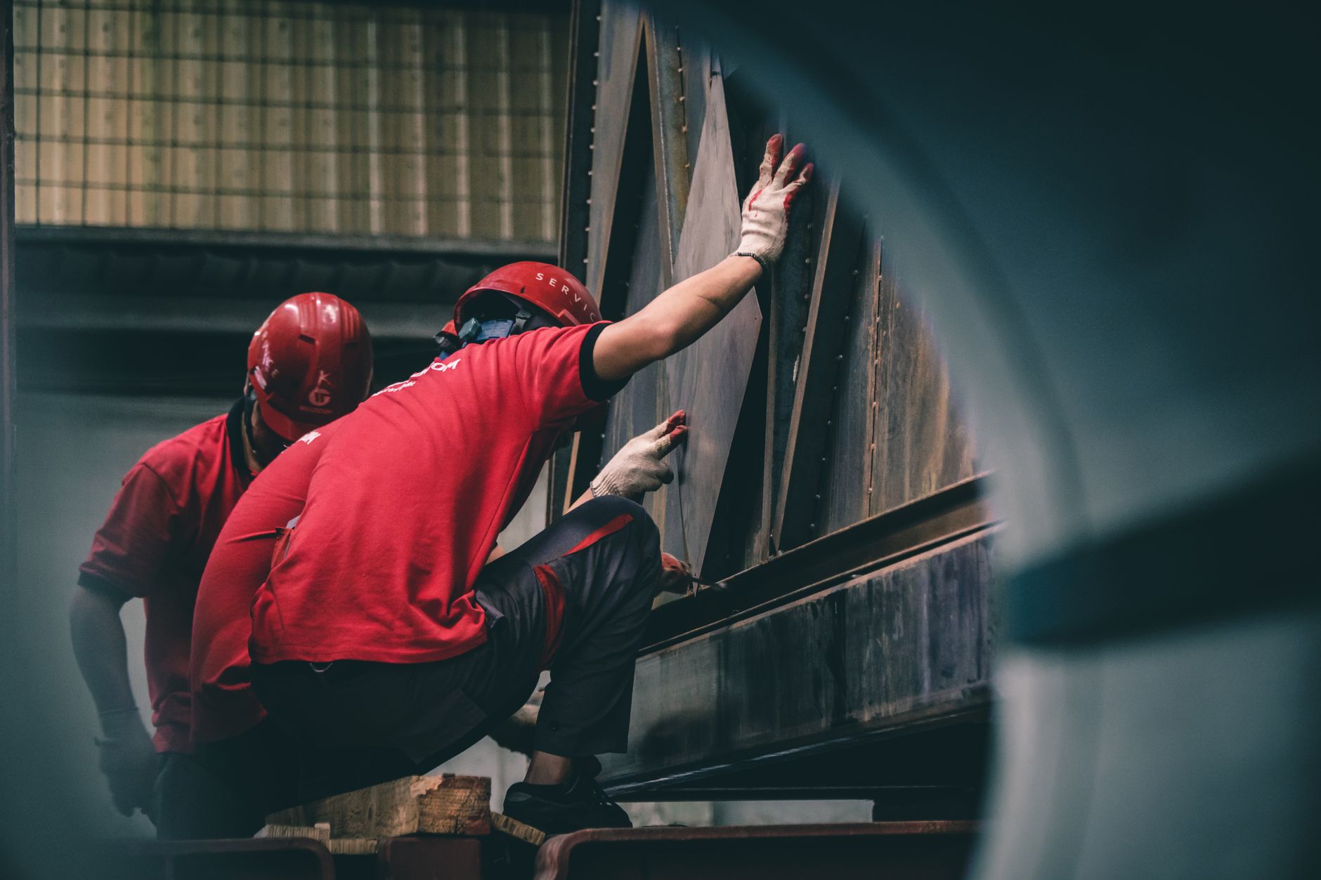 Dois trabalhadores usando capacetes vermelhos montam componentes metálicos dentro de uma fábrica.