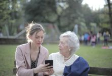 Mulher orienta uma idosa no uso do celular enquanto estão sentadas em um banco de parque.