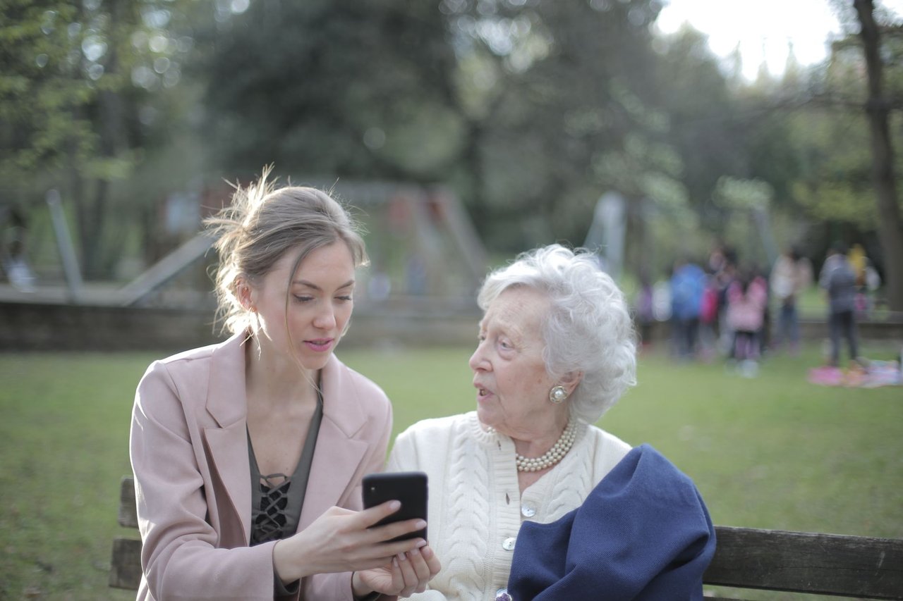 Mulher orienta uma idosa no uso do celular enquanto estão sentadas em um banco de parque.