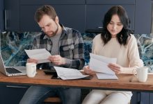 Casal analisa documentos financeiros em casa, usando um celular e conversando sobre o orçamento doméstico.