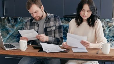 Casal analisa documentos financeiros em casa, usando um celular e conversando sobre o orçamento doméstico.